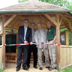Vier Männer stehen zur Einweihung in einem neuen, offenen Holzpavillon, dem Bienenschaustand. Gemeinsam durchschneiden sie ein rot-weißes Absperrband, um den Stand offiziell zu eröffnen.