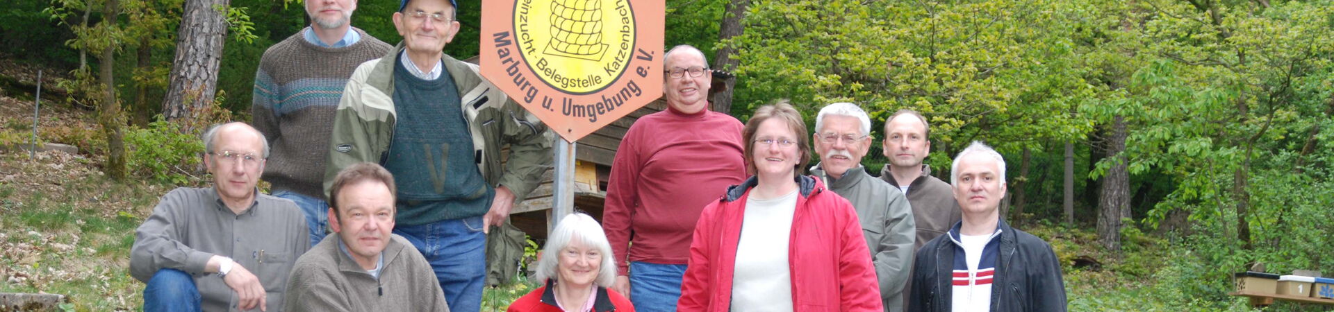 Zehn Mitglieder des Imkervereins Marburg und Umgebung posieren für ein Gruppenfoto im Freien. Sie stehen und sitzen auf einer grasbewachsenen Anhöhe mit Stufen. In ihrer Mitte ist ein Schild mit dem Vereinsnamen und -logo, einer Biene auf einer Wabe, zu s