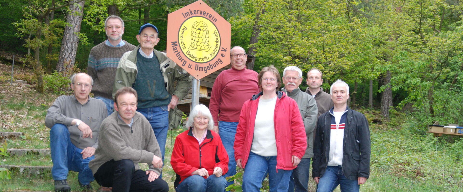 Zehn Mitglieder des Imkervereins Marburg und Umgebung posieren für ein Gruppenfoto im Freien. Sie stehen und sitzen auf einer grasbewachsenen Anhöhe mit Stufen. In ihrer Mitte ist ein Schild mit dem Vereinsnamen und -logo, einer Biene auf einer Wabe, zu s