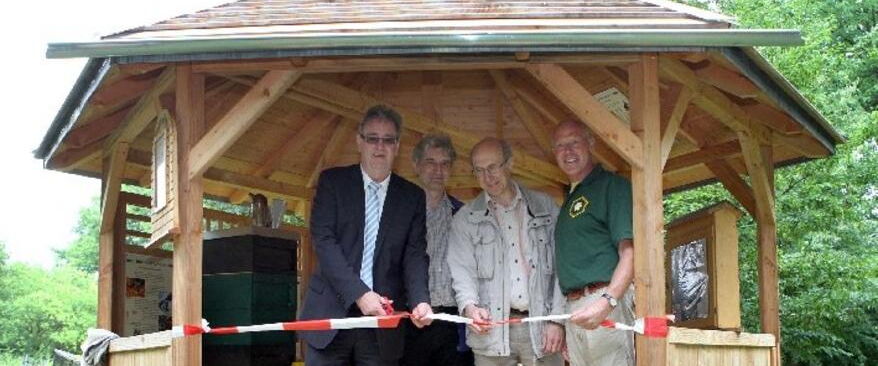 Vier Männer stehen zur Einweihung in einem neuen, offenen Holzpavillon, dem Bienenschaustand. Gemeinsam durchschneiden sie ein rot-weißes Absperrband, um den Stand offiziell zu eröffnen.